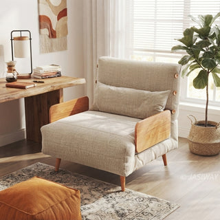 A beige linen convertible chair bed with wooden armrests and tapered legs sits on a patterned area rug in a sunlit room. A matching lumbar pillow is placed neatly on the seat. In the background, a live-edge wooden desk holds a lamp, coffee carafe, and books, next to a window with blinds and a potted plant in a woven basket.