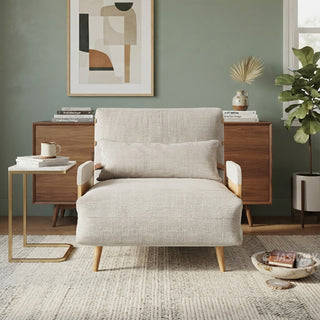 A beige linen convertible armchair with wooden armrests and tapered legs is centered on a textured area rug. A matching rectangular lumbar pillow rests against the backrest. In the background, a wooden sideboard features a framed abstract painting, books, and a vase, accompanied by a small side table and a potted plant.