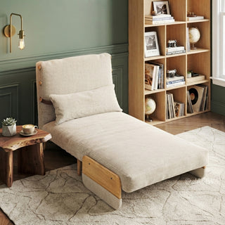 A beige linen convertible armchair is set up as an extended lounge chaise on a light patterned area rug. A matching rectangular lumbar pillow rests against its reclined backrest. In the background, a wooden bookshelf filled with books and decor sits next to a green-paneled wall, complemented by a live-edge side table and a wall sconce.