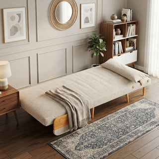 A fully flattened beige linen convertible chair bed rests on a wooden floor next to a patterned runner rug. A matching bolster pillow is placed at the head of the bed, and a textured throw blanket is draped across the middle. In the background, paneled walls feature framed art and a round mirror, alongside a bookshelf, a side table with a lamp, and a potted plant.