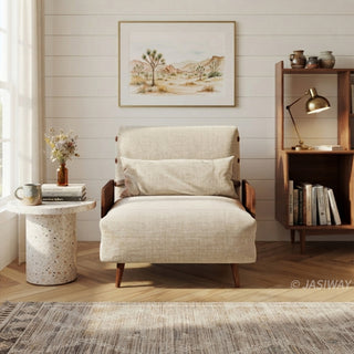 A beige linen convertible armchair with wooden armrests and legs sits on a patterned area rug in a bright room. A matching rectangular lumbar pillow rests against its backrest. In the background, a white shiplap wall features framed desert art, flanked by a terrazzo side table with flowers and a wooden bookshelf with a lamp.