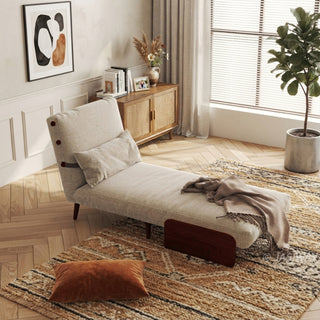 A beige linen convertible armchair set up as an extended lounge chaise rests on a patterned area rug in a sunlit room. A matching rectangular lumbar pillow leans against its reclined backrest, and a soft throw blanket drapes across the seat. In the background, a woven cabinet, framed wall art, and a potted plant complete the cozy interior.