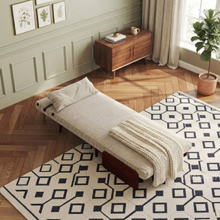 A fully flattened beige linen convertible chair bed rests on a geometric patterned area rug over a herringbone wooden floor. A matching pillow sits neatly at the head of the bed, and a textured cream throw blanket is draped across the middle. In the background, sage green paneled walls feature botanical framed art, a wooden sideboard with mugs, curtains, and a potted plant by the window.