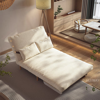 A beige linen convertible sofa bed is set up as an extended lounge chaise on a striped area rug in a sunlit room. Two matching rectangular lumbar pillows rest against its reclined backrest, and a floor lamp stands gracefully beside it. In the background, a wooden platform features a tea table, low seating, and built-in cabinetry, creating a warm and inviting atmosphere.