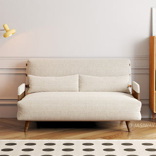 A beige linen convertible loveseat sits against a paneled wall over a herringbone wooden floor and a dotted area rug. Two matching long bolster pillows are placed neatly against its supportive backrest. A yellow wall sconce illuminates the left side, while a wooden cabinet stands on the right to complete the warm living room setting.
