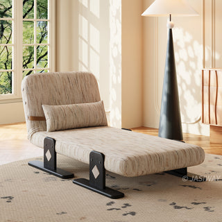 A modern low-profile lounge chair with beige textured fabric and a matching lumbar pillow rests on a patterned rug. The chair features dark wooden legs with unique diamond-shaped accents and stands near a large window. In the background, a tall, conical floor lamp with a white shade casts a warm glow against the pale walls.