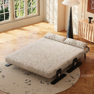 A high-angle shot shows a modern, wide beige textured lounger fully reclined flat into a bed configuration. The low-profile furniture rests on a round patterned rug over a herringbone wooden floor. Two matching long cylindrical pillows are positioned at the head of the flat surface. In the background, a tall dark conical floor lamp and a wooden cabinet stand near a sunlit window.