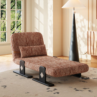A modern low-profile lounge chair upholstered in rich rust-brown textured fabric sits on a light patterned rug. The chair features a matching rectangular lumbar pillow and is supported by dark wooden block legs with light diamond accents. In the background, a tall conical floor lamp with a white shade stands beside a sunlit window and wooden cabinet.