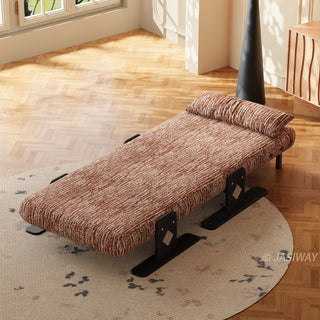An overhead, angled view shows a modern rust-brown textured lounger fully reclined into a flat bed position. The low-profile piece rests on a round patterned rug over a herringbone wood floor, accompanied by a matching long bolster pillow. In the background, a tall black conical floor lamp and a wooden cabinet stand near a sunlit window.