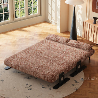 An elevated angle showcases a modern, wide rust-brown textured lounger fully unfolded into a flat bed configuration. The low-profile piece rests on a round patterned rug atop a herringbone wood floor, complete with two matching bolster pillows. In the background, a tall black conical lamp and a wooden cabinet stand near a sunlit window.