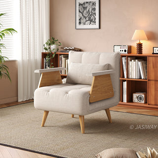 A beige tufted fabric armchair with wooden legs and distinctive angled armrests sits on a textured area rug in a cozy room. A matching rectangular lumbar pillow is neatly arranged against the backrest. In the background, a warm wooden bookshelf filled with books, a small lamp, and a framed botanical artwork complete the inviting interior.