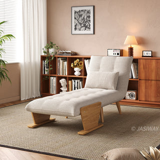 A beige tufted fabric convertible sofa is set up as a lounge chaise on a textured area rug in a sunlit room. A matching rectangular lumbar pillow rests against its reclined backrest. In the background, a warm wooden bookcase filled with books, a small table lamp, and framed wall art complete the cozy interior.