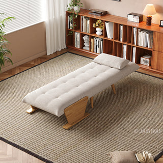 A fully flattened beige tufted fabric convertible sofa bed rests on a textured area rug in a bright room. A matching pillow is placed neatly at the head of the bed. In the background, a long wooden bookshelf filled with books, a window with blinds, and a small table lamp complete the cozy setup.