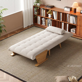 A modern convertible daybed fully extended into a flat sleeping position is upholstered in textured beige fabric and sits on a neutral woven area rug over a wooden floor. The mattress surface features neat button-tufting, accompanied by a matching rectangular pillow and distinct wooden side panels that act as a sturdy base. In the background, a cozy room setting with a long wooden bookshelf filled with books, a small table lamp, and a window with white blinds completes the warm, inviting aesthetic.
