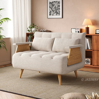 A wide beige tufted fabric loveseat with wooden legs and distinctive wooden armrests sits on a textured area rug in a sunlit living room. Two matching rectangular lumbar pillows are neatly arranged against the backrest. In the background, a warm wooden bookcase filled with books, a small table lamp, and framed wall art complete the cozy interior.