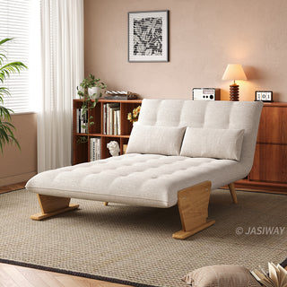 A beige tufted fabric convertible sofa is set up as a lounge chaise on a textured area rug in a sunlit room. Two matching rectangular lumbar pillows rest against its reclined backrest. In the background, a warm wooden bookcase filled with books, a small table lamp, and framed wall art complete the cozy interior.
