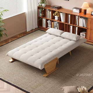 A fully flattened beige tufted fabric convertible sofa bed rests on a textured area rug in a bright room. Two matching pillows are placed neatly at the head of the bed. In the background, a long wooden bookshelf filled with books, a window with blinds, and a small table lamp complete the cozy setup.