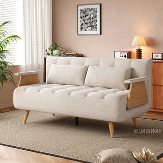 A wide beige tufted fabric loveseat with wooden legs and distinctive wooden armrests sits on a textured area rug in a sunlit living room. Two matching rectangular lumbar pillows are neatly arranged against the backrest. In the background, a warm wooden cabinet, a table lamp, and framed wall art complete the cozy interior.