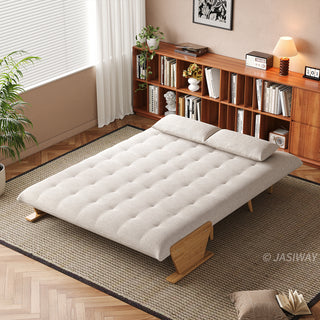 A fully flattened beige tufted fabric convertible sofa bed rests on a textured area rug in a bright room. Two matching pillows are placed neatly at the head of the bed. In the background, a long wooden bookshelf filled with books, a window with blinds, and a small table lamp complete the cozy setup.
