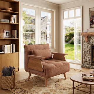 A rust-brown tufted fabric armchair with wooden legs and distinctive wooden armrests sits on a patterned area rug in a sunlit living room. A matching rectangular lumbar pillow is neatly arranged against the backrest. In the background, large windows and an open door reveal a green garden view, complemented by a tall wooden bookcase and a stone fireplace.