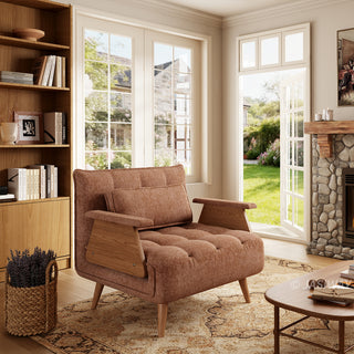 A modern convertible armchair upholstered in textured brown fabric sits on a patterned area rug over a wooden floor. The armchair features button-tufting on the seat and back cushions, a matching rectangular lumbar pillow, wooden armrest panels, and tapered wooden legs. In the background, a cozy sunlit room with a built-in bookshelf, a stone fireplace, and open glass doors overlooking a lush green garden creates a warm and inviting atmosphere.