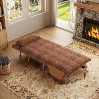 A fully flattened rust-brown tufted fabric convertible sofa bed rests on a patterned area rug in a cozy living room. A matching pillow is positioned neatly at the head of the bed. In the background, open glass doors reveal a green garden view next to a warm stone fireplace, completing the inviting interior.