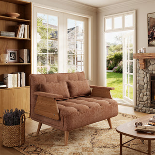 A rust-brown tufted fabric loveseat with wooden legs and distinctive wooden armrests sits on a patterned area rug in a sunlit living room. Two matching rectangular lumbar pillows are neatly arranged against the backrest. In the background, open glass doors reveal a green garden view, complemented by a tall wooden bookcase, a coffee table, and a stone fireplace.
