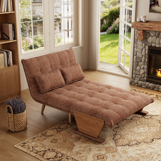 A rust-brown tufted fabric convertible sofa is set up as a lounge chaise on a patterned area rug in a sunlit room. Two matching rectangular lumbar pillows rest against its reclined backrest. In the background, open glass doors reveal a green garden view next to a warm stone fireplace, completing the inviting interior.