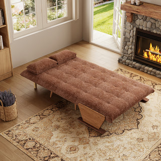 A fully flattened rust-brown tufted fabric convertible sofa bed rests on a patterned area rug in a cozy living room. Two matching pillows are positioned neatly at the head of the bed. In the background, open glass doors reveal a green garden view next to a warm stone fireplace, completing the inviting interior.