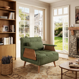 A sage green tufted fabric armchair with wooden legs and distinctive wooden armrests sits on a patterned area rug in a sunlit living room. A matching rectangular lumbar pillow is neatly arranged against the backrest. In the background, large windows open to a green garden view, complemented by a tall wooden bookcase and a stone fireplace.