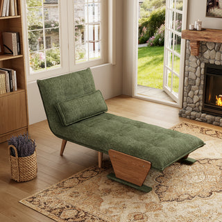 A sage green tufted fabric convertible sofa is set up as a lounge chaise on a patterned area rug in a cozy, sunlit room. A matching rectangular lumbar pillow rests against its reclined backrest. In the background, open glass doors reveal a green garden view, complemented by a stone fireplace, a basket of lavender, and a wooden bookshelf.