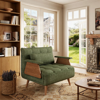 A modern convertible armchair upholstered in textured green fabric sits on a patterned area rug over a wooden floor. The armchair features button-tufting on the seat and back cushions, a matching rectangular lumbar pillow, wooden armrest panels, and tapered wooden legs. In the background, a cozy sunlit room with a built-in bookshelf, a stone fireplace, and open glass doors overlooking a lush green garden creates a warm and inviting atmosphere.