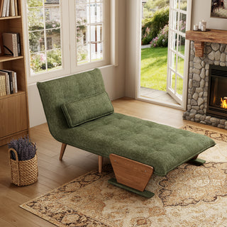 A modern convertible chaise lounge upholstered in textured green fabric is set up as a recliner on a patterned area rug over a wooden floor. The lounge features an adjustable backrest with button-tufting, a matching rectangular bolster pillow, and wooden side supports combined with tapered wooden legs. In the background, a cozy room with a built-in bookshelf, a stone fireplace, and open glass doors overlooking a sunny garden creates a warm and inviting atmosphere.