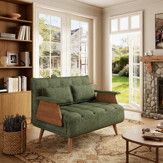 A sage green tufted fabric loveseat with wooden legs and distinctive wooden armrests sits on a patterned area rug in a sunlit living room. Two matching rectangular lumbar pillows are neatly arranged against the backrest. In the background, open glass doors reveal a green garden view, complemented by a tall wooden bookcase, a coffee table, and a stone fireplace.