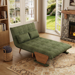 A sage green tufted fabric convertible sofa is set up as a lounge chaise on a patterned area rug in a sunlit room. Two matching rectangular lumbar pillows rest against its reclined backrest. In the background, open glass doors reveal a green garden view next to a warm stone fireplace, completing the inviting interior.
