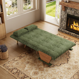 A fully flattened sage green tufted fabric convertible sofa bed rests on a patterned area rug in a cozy living room. Two matching pillows are positioned neatly at the head of the bed. In the background, open glass doors reveal a green garden view next to a warm stone fireplace, completing the inviting interior.