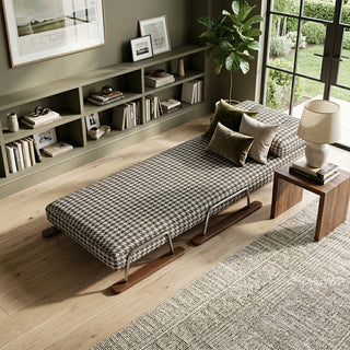 A black and white houndstooth pattern convertible sleeper sofa fully extended into a single bed, styled with olive green accent pillows in a sophisticated living room with built-in dark shelving, hardwood flooring, and a large side table with a lamp.