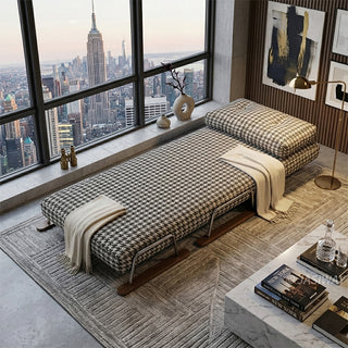 A black and white houndstooth pattern convertible sleeper sofa fully extended into a single bed in a luxury New York high-rise apartment, featuring floor-to-ceiling city skyline views, a draped cream throw blanket, an area rug, and contemporary wall art.