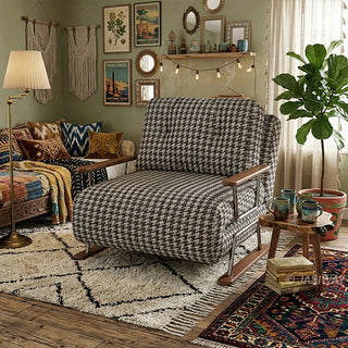 A black and white houndstooth pattern convertible sleeper sofa armchair featuring metal and wood-trimmed arms in a boho-style living room with macramé wall hangings, a gallery wall, string lights, layered vintage rugs, and a potted fiddle-leaf fig tree.