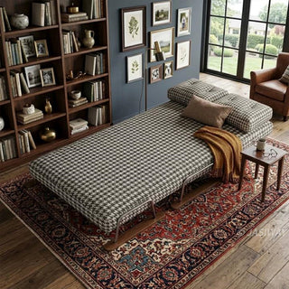 A brown and white houndstooth pattern convertible sleeper sofa fully extended into a single bed, styled with a gold throw blanket and accent pillows in a cozy living room featuring a large tall bookcase, hardwood floors, and a vintage patterned rug.