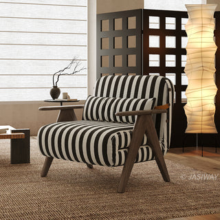 A black-and-white striped convertible armchair sits on a textured brown rug in a softly lit modern room. A matching striped lumbar pillow rests against its backrest, framed by dark wooden armrests and legs. In the background, a warm vertical paper floor lamp, a decorative screen, and a vase with dry branches complete the cozy interior staging.