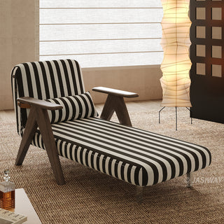 A black-and-white striped convertible armchair is set up as an extended lounge chaise on a textured brown rug in a softly lit room. A matching striped lumbar pillow rests against its backrest, framed by dark wooden armrests and V-shaped legs. In the background, a warm vertical paper floor lamp and a decorative screen complete the cozy interior staging.