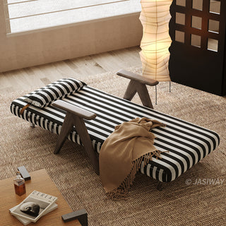 A black-and-white striped convertible armchair is fully flattened into a sleek single bed on a textured brown rug in a softly lit room. A matching striped pillow rests at the head of the bed, while a warm brown throw blanket drapes across the middle. In the background, a warm vertical paper floor lamp and a dark wooden screen complete the cozy interior staging.