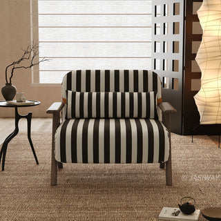 A black-and-white striped convertible armchair sits centered on a textured brown rug in a warmly lit modern room. A matching striped lumbar pillow is positioned neatly against its backrest, framed by wooden armrests and legs. In the background, a small side table with a vase, a tall vertical paper floor lamp, and a decorative screen complete the cozy interior staging.
