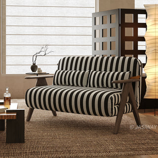 A black-and-white striped convertible loveseat sits on a textured brown rug in a softly lit modern room. Two matching striped pillows rest against its supportive backrest, flanked by dark wooden armrests and legs. In the background, a small side table with a vase, a tall vertical paper floor lamp, and a decorative screen complete the cozy interior staging.