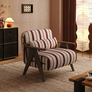 A red-and-white striped convertible armchair sits on a textured brown rug in a warmly lit modern room. A matching striped lumbar pillow rests against its backrest, framed by dark wooden armrests and legs. In the background, a tall vertical paper floor lamp, a dark storage cabinet with a small table lamp, and rich brown curtains complete the cozy interior staging.
