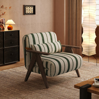 A green-and-white striped convertible armchair sits on a textured brown rug in a warmly lit modern room. A matching striped lumbar pillow rests against its backrest, framed by dark wooden armrests and legs. In the background, a tall vertical paper floor lamp, a dark storage cabinet with a small table lamp, and rich brown curtains complete the cozy interior staging.