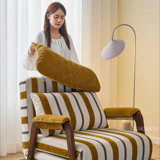 A model wearing a white blouse stands behind a yellow-and-white striped convertible armchair, adjusting its plush matching headrest cushion. Another matching striped lumbar pillow rests against the back of the chair, which features dark wooden armrests and legs. In the background, a modern floor lamp and white window blinds complete the bright interior scene.