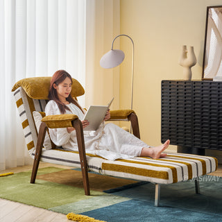 A model in a white dress lounges comfortably on an extended yellow-and-white striped convertible chaise chair while reading a book. The reclined furniture features a matching headrest cushion, a supportive lumbar pillow, and dark wooden armrests with sleek legs. She rests on a colorful block-patterned rug inside a brightly lit room with a modern floor lamp, a side cabinet, and window blinds in the background.