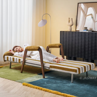 A model in a white dress takes a nap lying fully extended on a yellow-and-white striped convertible chaise lounge. The fully reclined furniture piece features a matching headrest cushion, dark wooden armrests, and sturdy legs supporting the extended frame. She rests peacefully on a colorful block-patterned rug in a brightly lit modern living room with a floor lamp and a dark cabinet in the background.